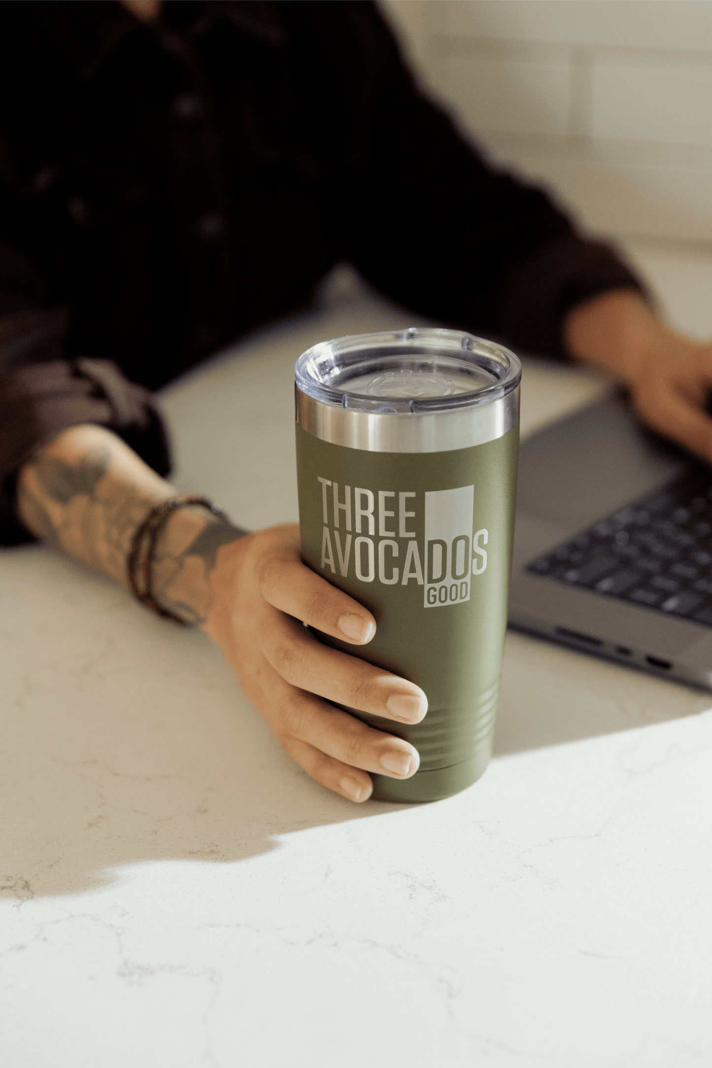 A hand holding olive green tumbler (20oz) engraved with Three Avocados logo.