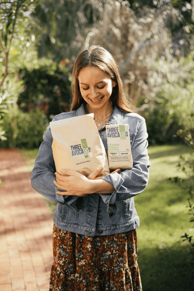 A woman smiling in the Sun holding a 12oz bag of coffee and a larger bulk bag