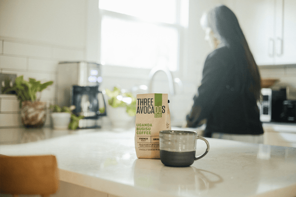 A woman washes dishes in the background while a bag of coffee and mug are in focus on the counter